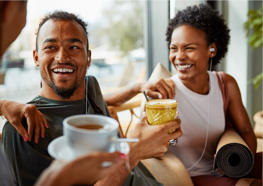 Two friends laughing at a cafe, wearing Nuropod earpieces and holding coffee and matcha drinks