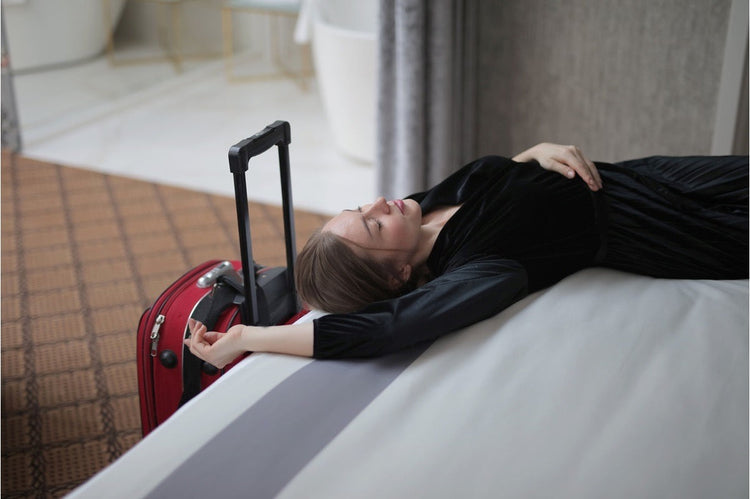 Woman lying across a hotel bed beside her suitcase, worn out after a long trip, the kind of travel strain that can lower HRV.